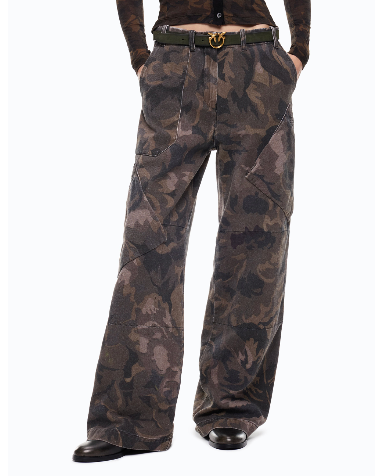 A person wearing wide-leg camouflage pants with a dark olive belt and black shoes, shown from the waist down against a plain white studio background.