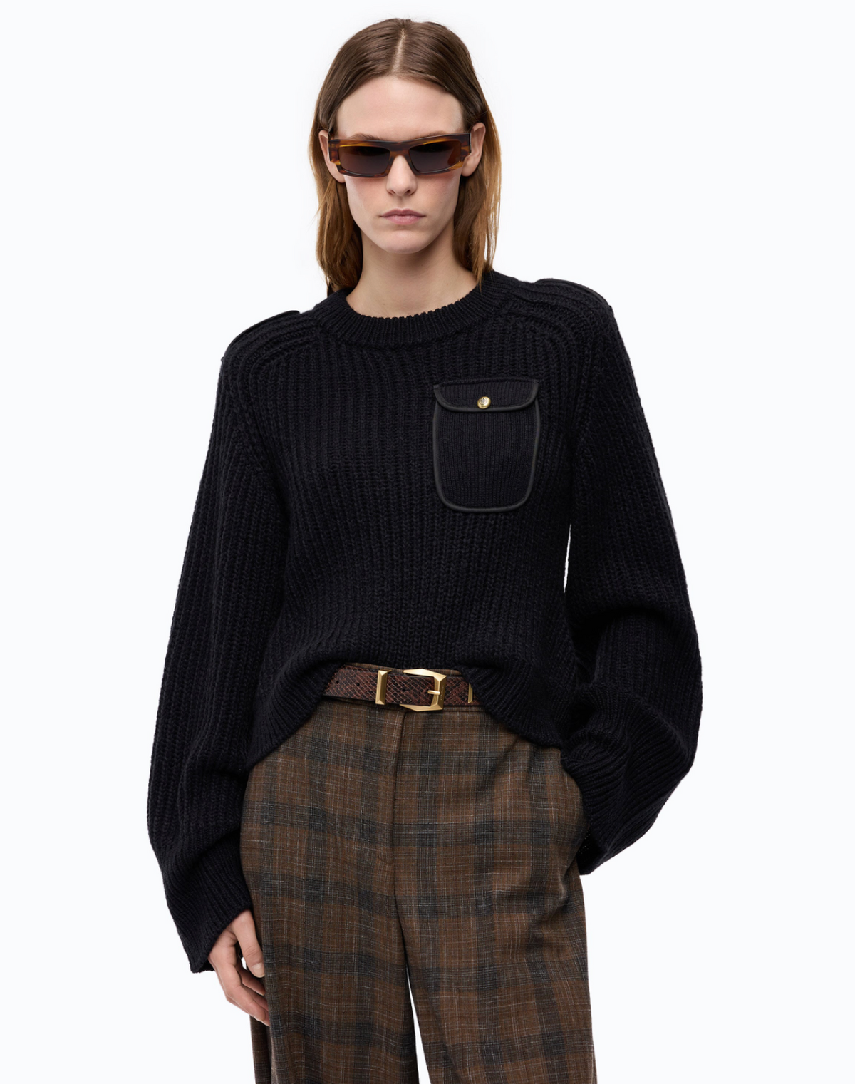 A model wearing a black ribbed knit crewneck sweater with a chest pocket, paired with brown plaid trousers and sunglasses, against a plain white background.