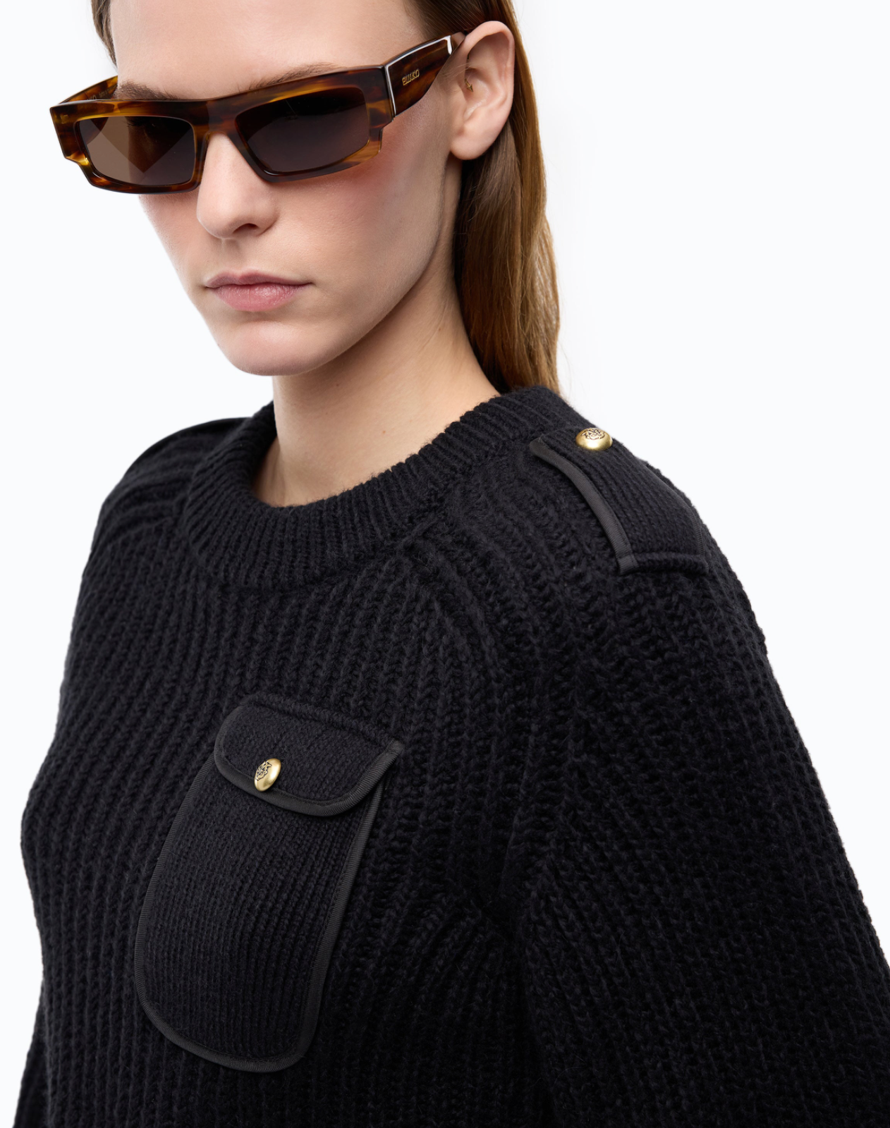 Close-up of a model wearing a black ribbed knit sweater with black leather-like shoulder tab details and a matching chest pocket, accented by gold-tone buttons. The model is also wearing tortoiseshell sunglasses against a plain white background.
