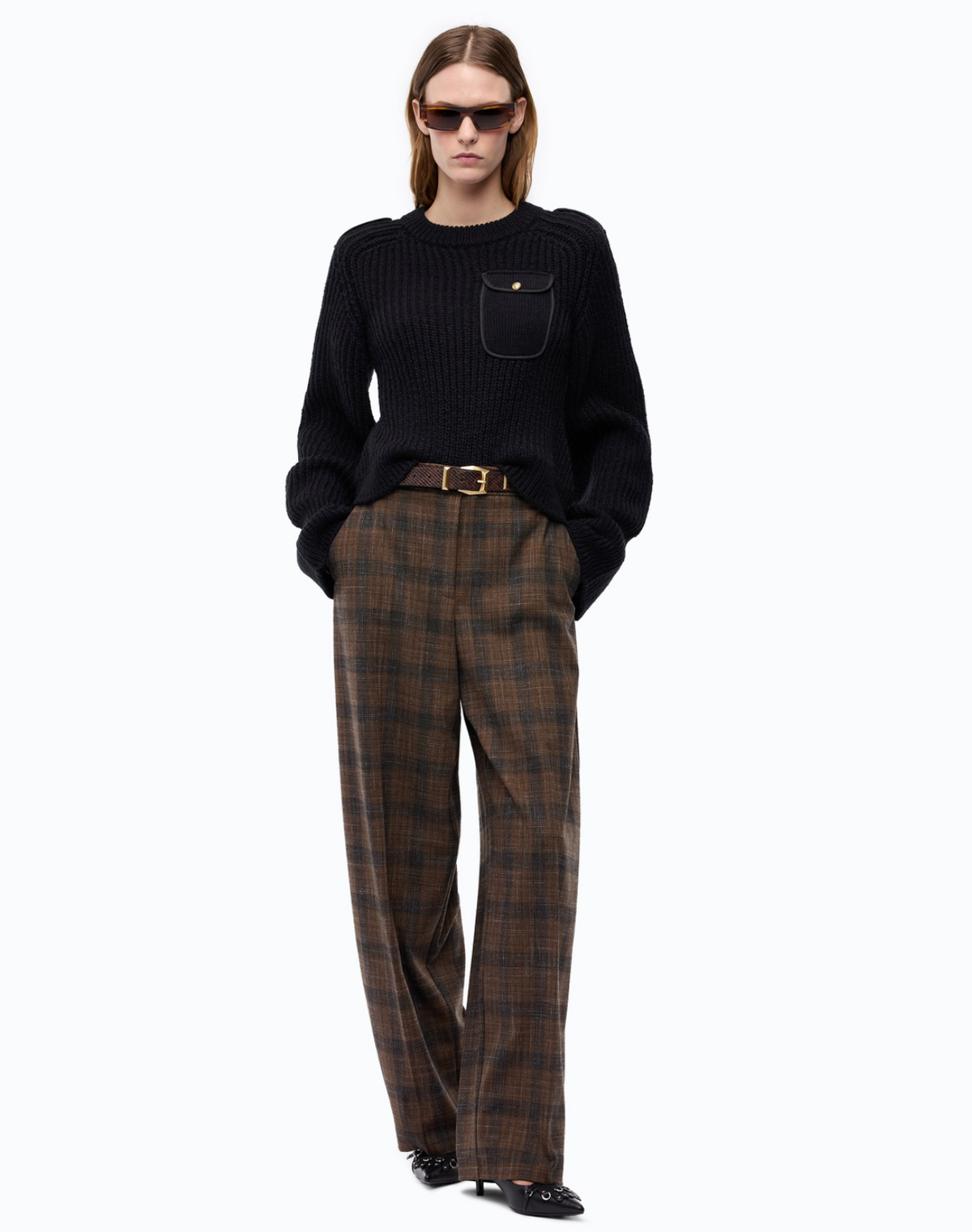 A model wearing a black ribbed knit sweater with a chest pocket, brown and black plaid trousers, a gold-brown belt, and black sunglasses, standing against a plain white background.