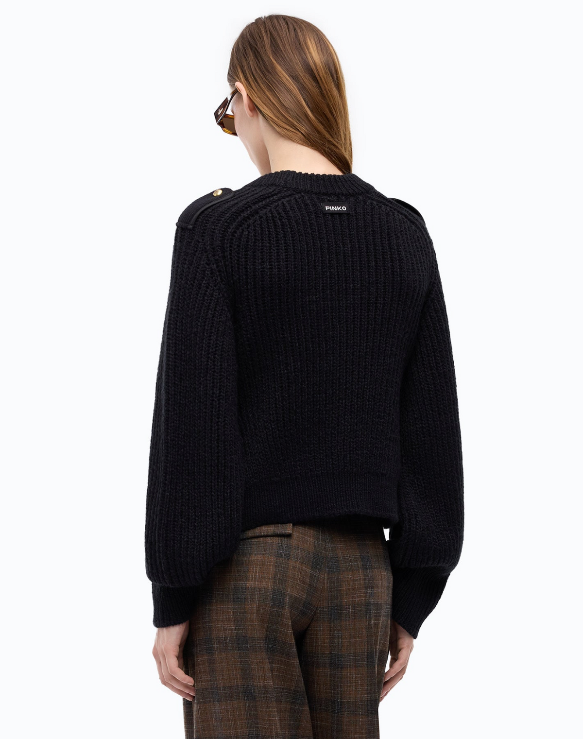 A model shown from the back wearing a black ribbed knit cropped sweater with shoulder details and brown plaid trousers, against a plain white background.