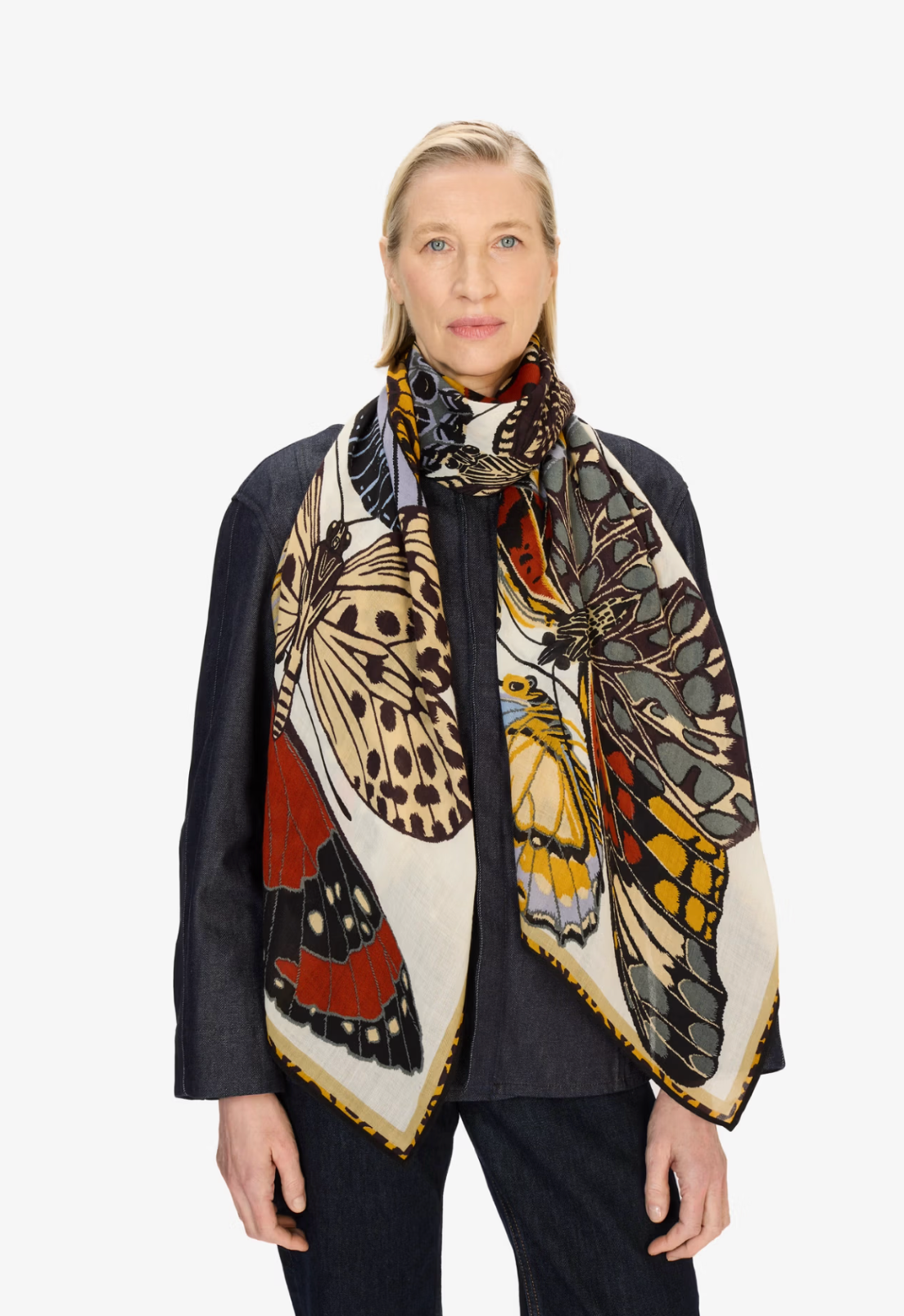 A woman modeling a large square scarf with a detailed butterfly and moth print in cream, black, orange, yellow, and red, styled over a dark navy jacket and jeans against a plain white studio background.