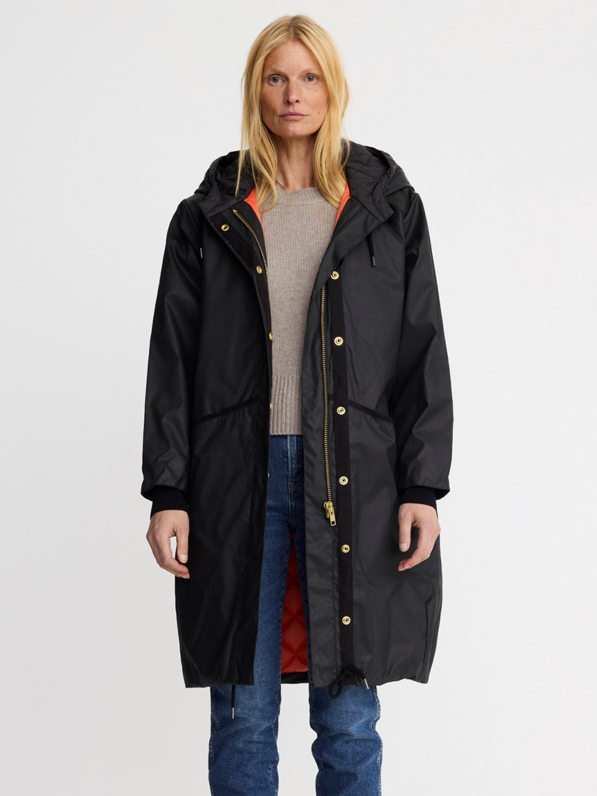 A woman modeling a long black hooded parka with gold-tone snap and zipper details, worn over a beige sweater and blue jeans against a plain light gray studio background.