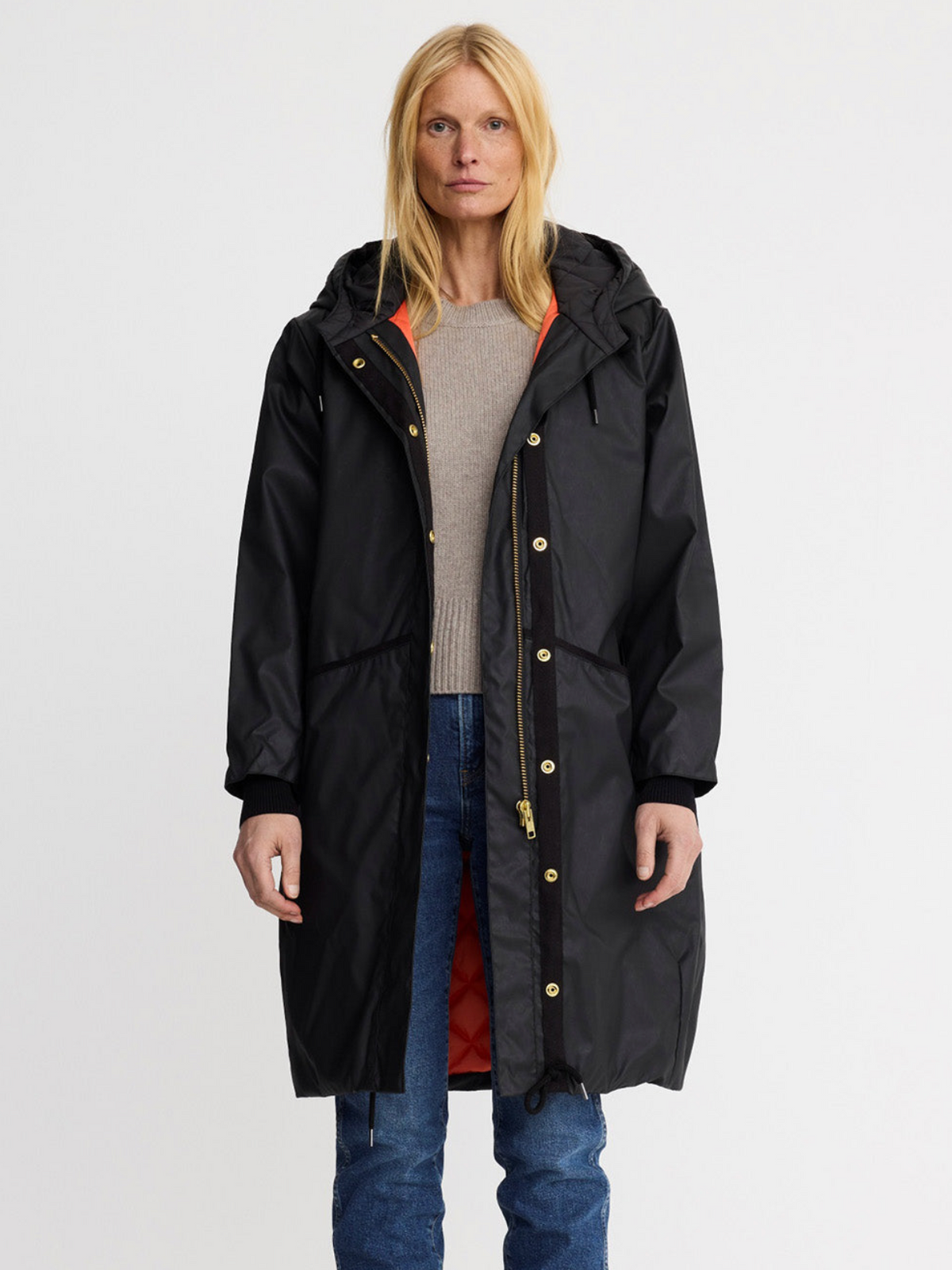 A woman modeling a long black hooded parka with gold-tone snap and zipper details, worn over a beige sweater and blue jeans against a plain light gray studio background.