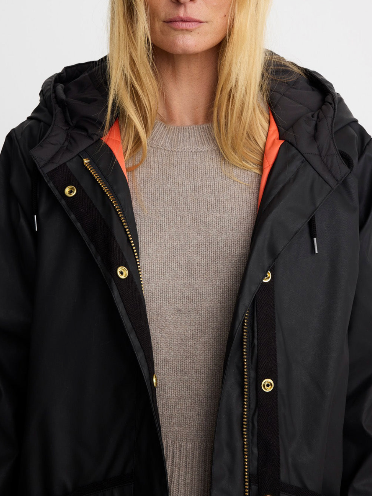 A woman modeling a long black hooded parka with gold-tone snap and zipper details, worn over a beige sweater and blue jeans against a plain light gray studio background.