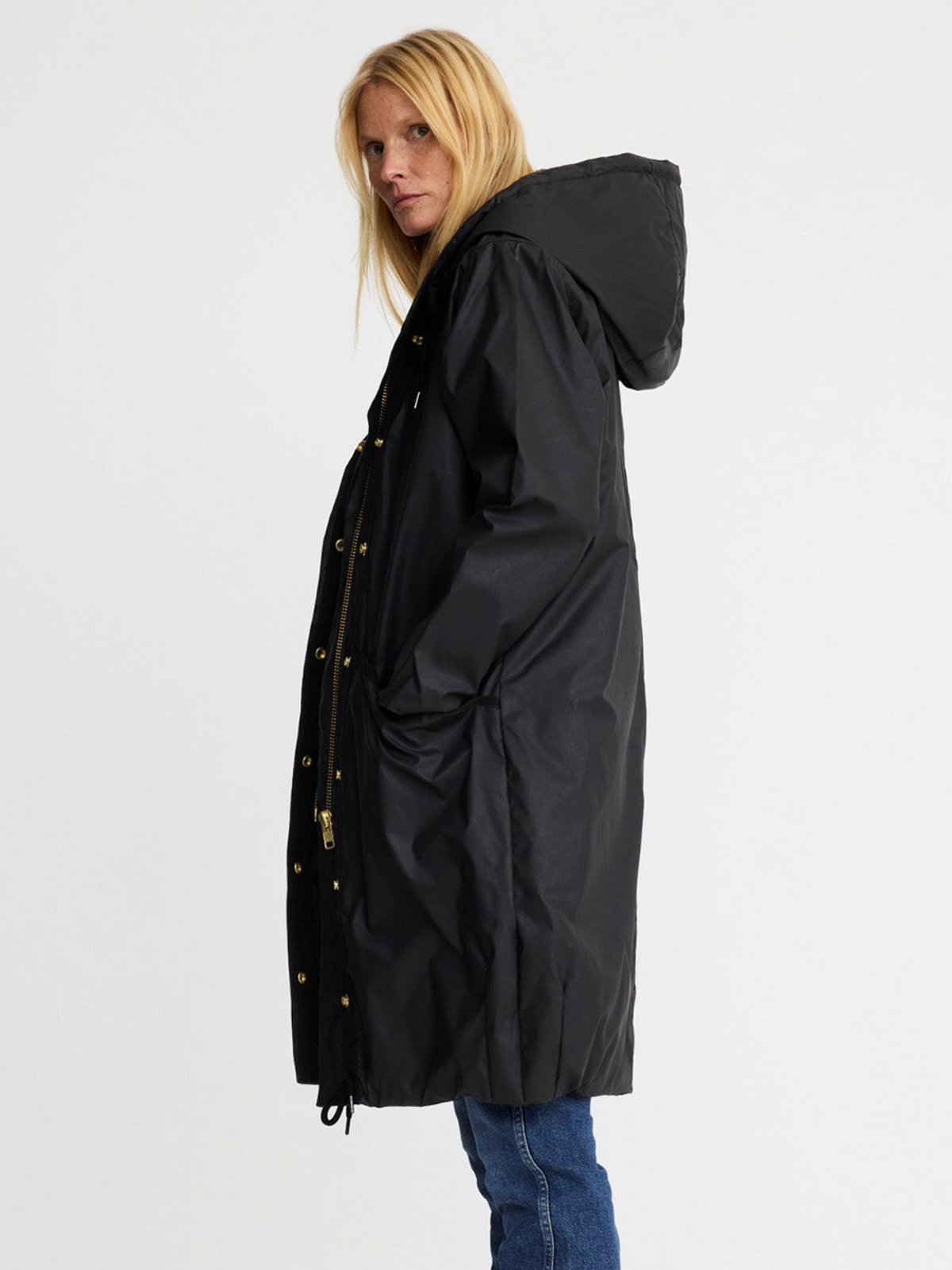 A woman wearing a long black hooded parka jacket with gold-tone snap and zipper details, paired with blue jeans, photographed against a plain light gray studio background.