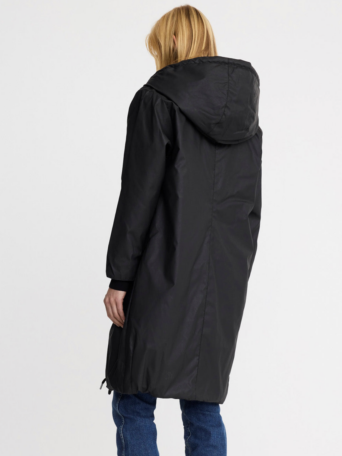 A woman viewed from the back wearing a long black hooded coat over blue jeans, photographed against a plain light gray studio background.