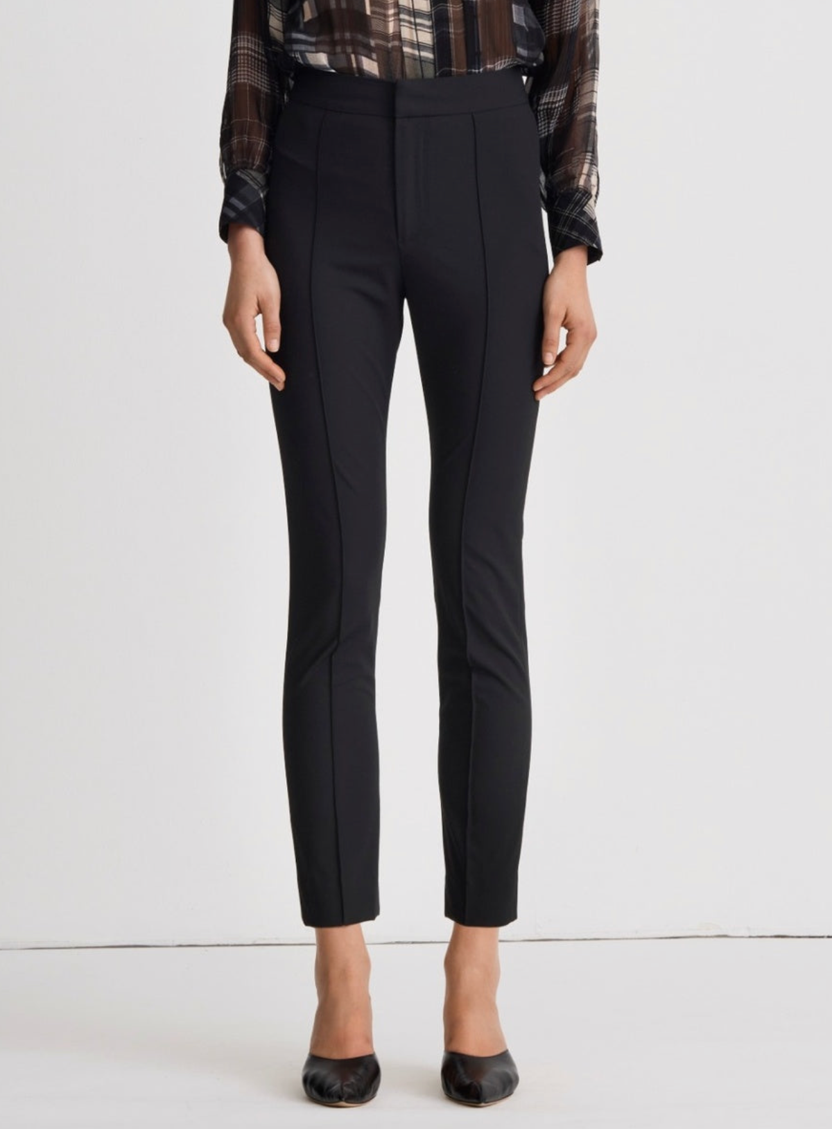 A model is shown from the waist down wearing black slim-fit ankle-length trousers, a brown and black plaid shirt, and black pointed-toe heels against a plain white studio background.