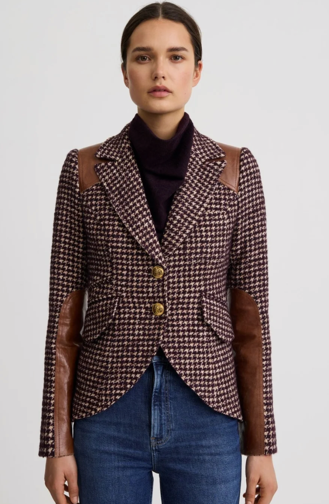 A model wearing a brown and black houndstooth blazer with brown faux-leather elbow patches and gold buttons, styled with a dark turtleneck and blue jeans against a light gray background.