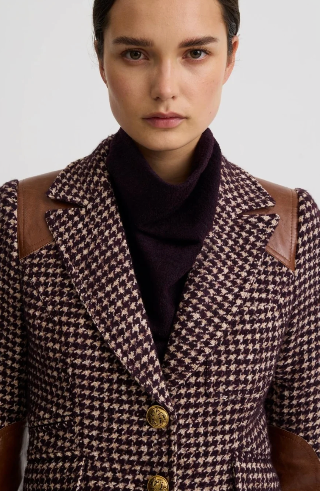A model wearing a brown and black houndstooth blazer with brown faux-leather elbow patches and gold buttons, styled with a dark turtleneck and blue jeans against a light gray background.