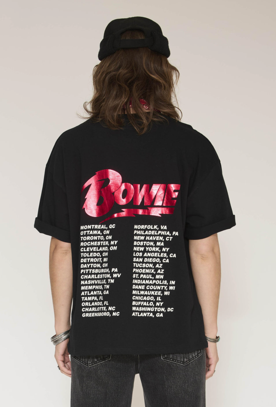 A person shown from behind wearing a black oversized T-shirt with a large pink and red 'Rome' graphic across the back and a list of city names in white text below, paired with a black beanie and dark jeans against a plain light background.