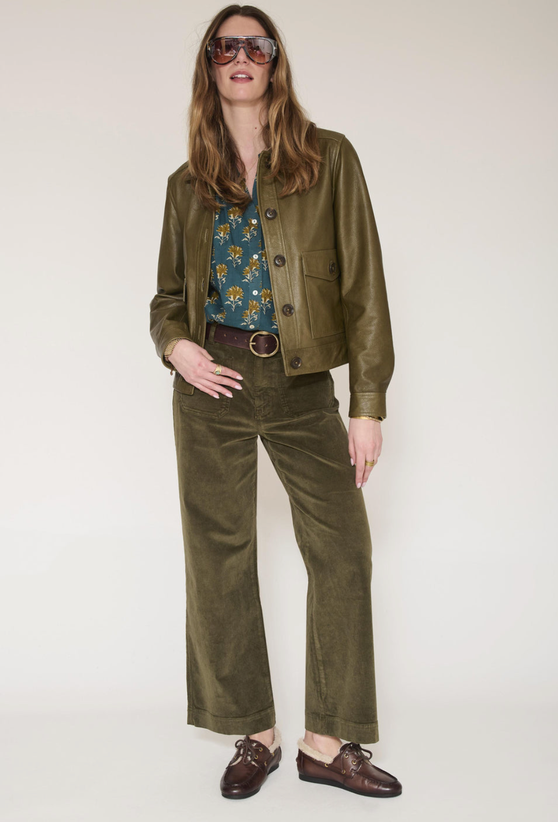 A woman modeling an olive green corduroy jacket and matching wide-leg trousers, styled with a floral blue top, brown leather loafers, and sunglasses against a plain light background.