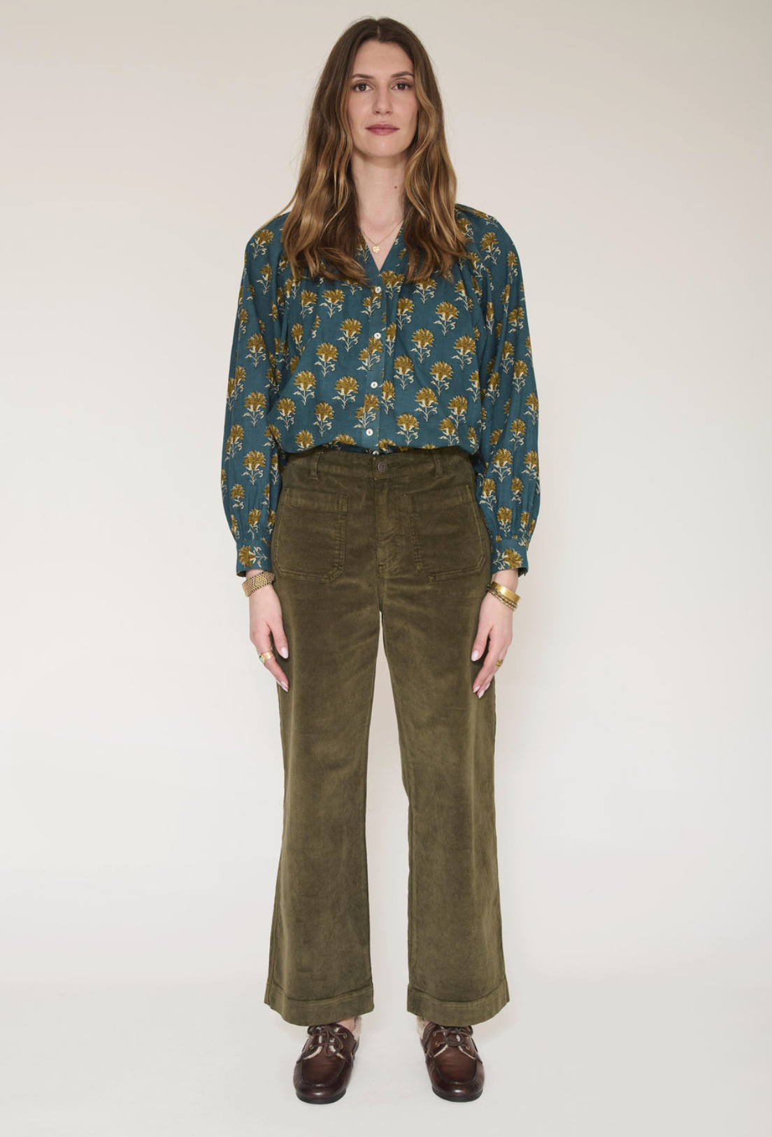 A woman modeling an olive green corduroy jacket and matching wide-leg trousers, styled with a floral blue top, brown leather loafers, and sunglasses against a plain light background.