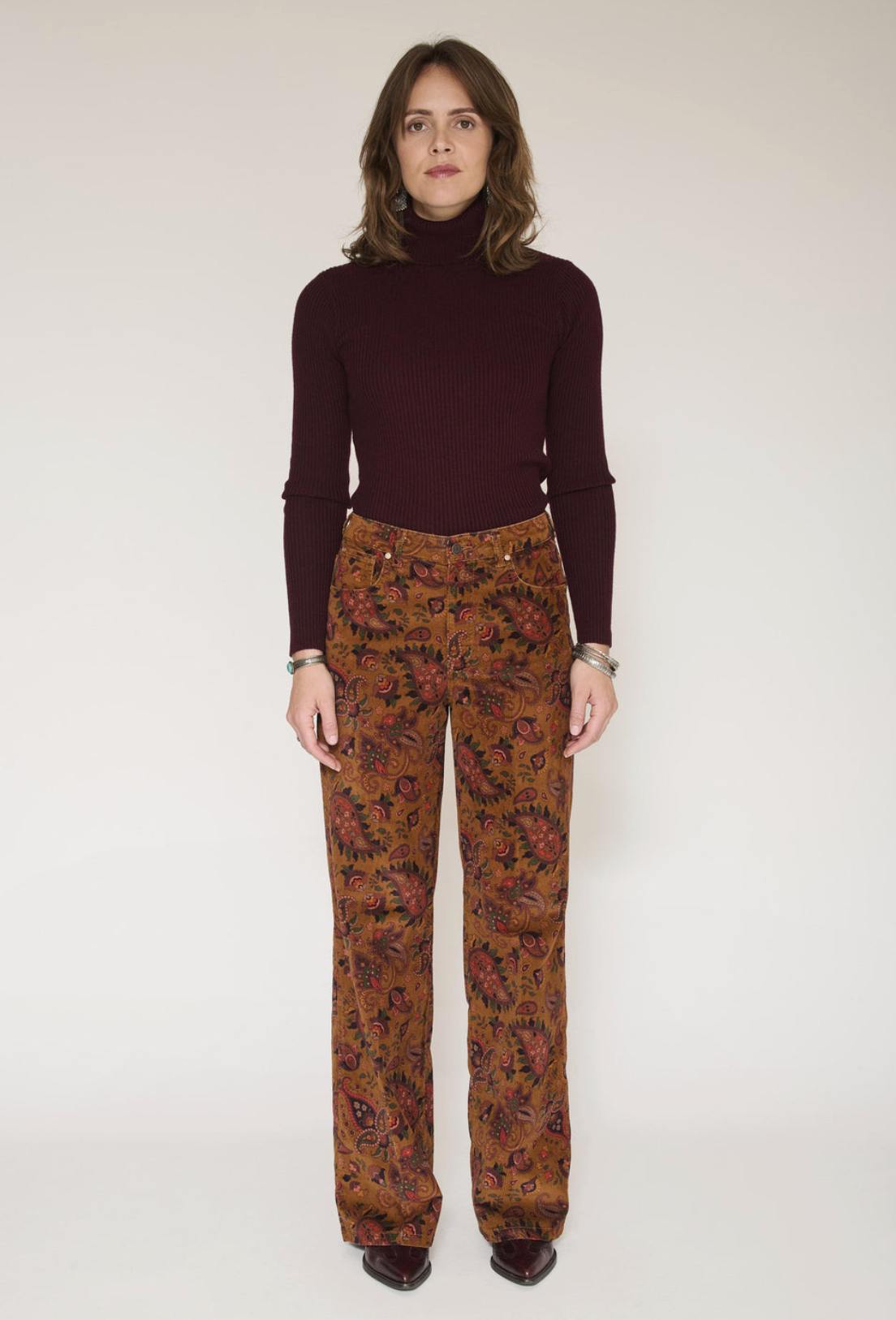 A model stands against a plain light beige studio background wearing a dark burgundy ribbed turtleneck sweater and brown wide-leg trousers with a dense multicolor floral pattern.