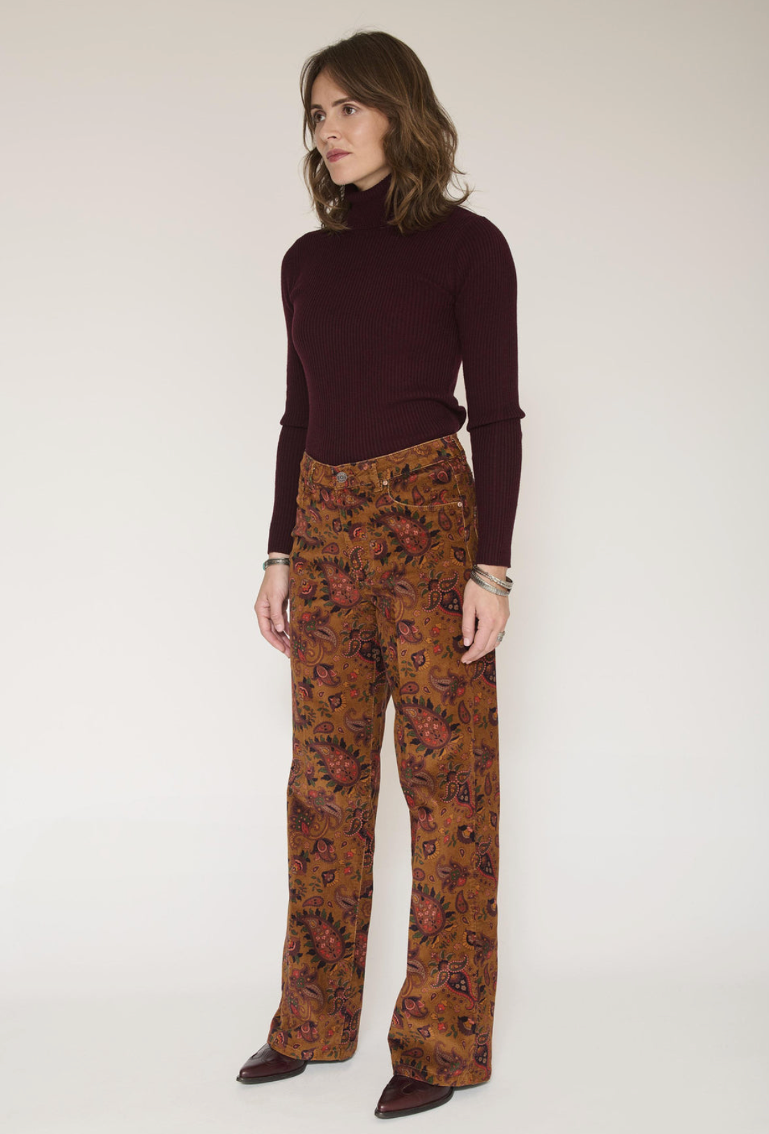 A model stands against a plain light beige studio background wearing a dark burgundy ribbed turtleneck sweater and brown wide-leg trousers with a dense multicolor floral pattern.