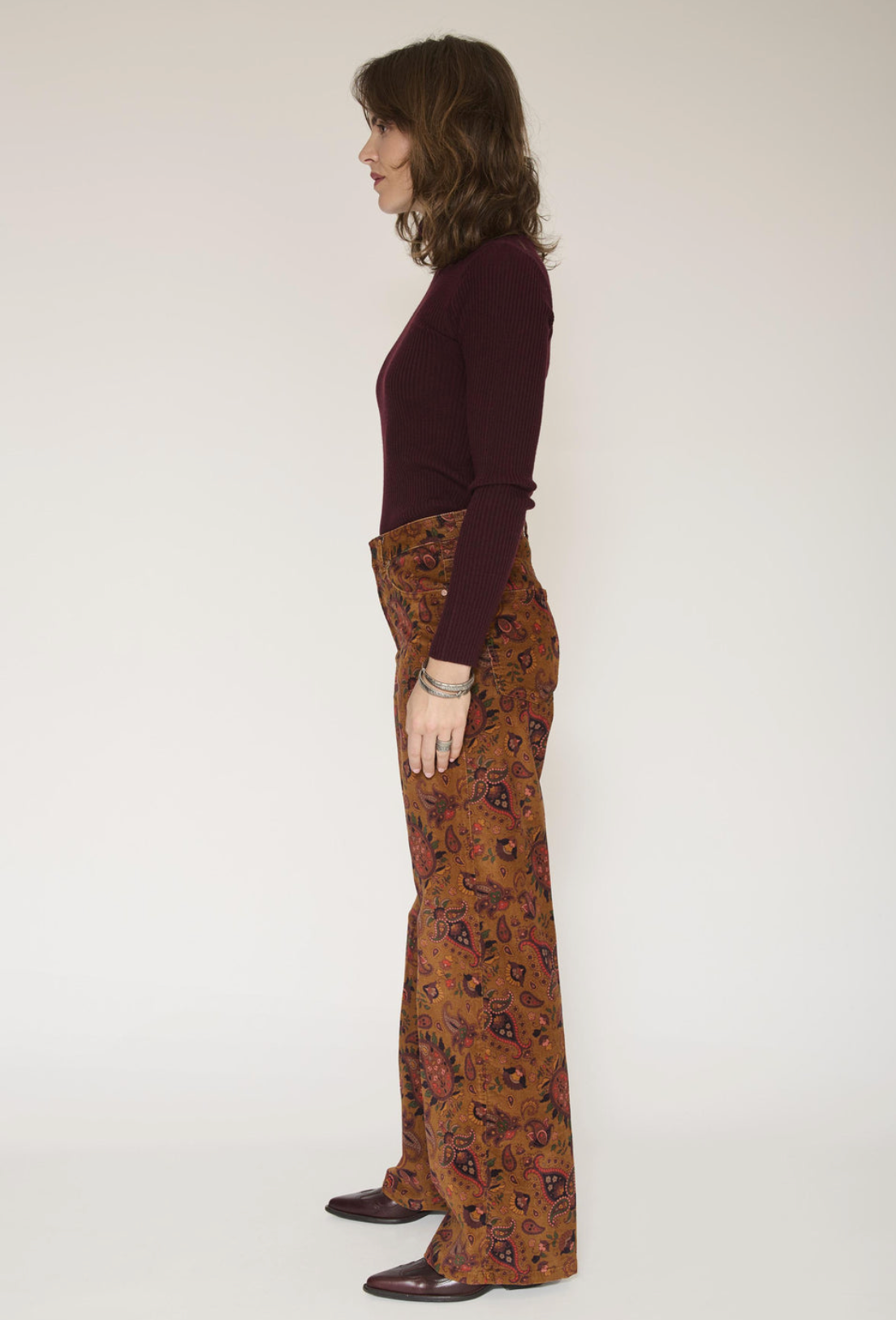 A woman stands in profile wearing a dark burgundy long-sleeve top and wide-leg brown pants with a red, black, and gold paisley pattern, paired with dark brown shoes, against a plain off-white studio background.