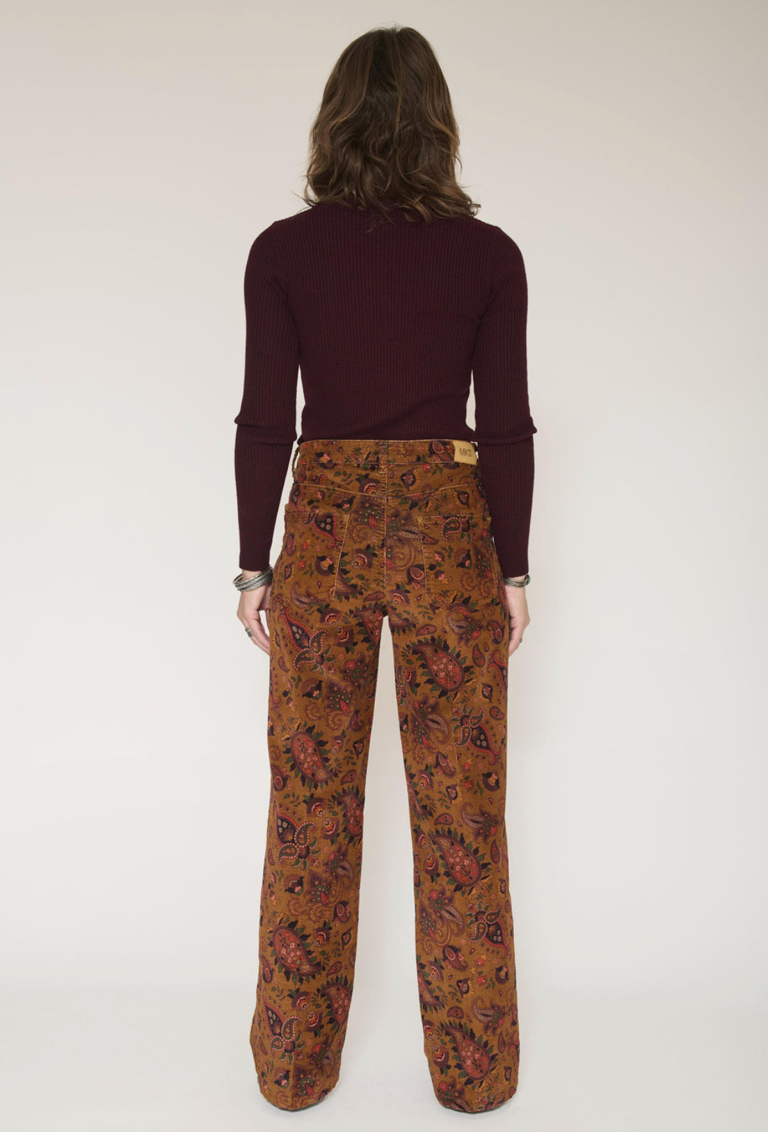 A model viewed from the back wearing a dark burgundy long-sleeve top and brown wide-leg trousers with a red and black floral pattern, set against a plain off-white studio background.