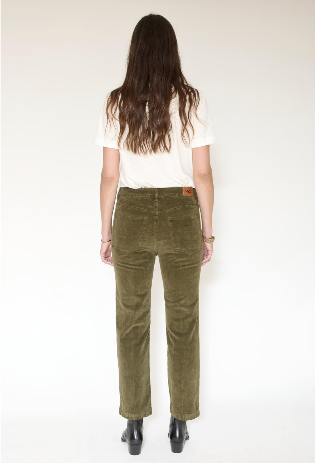 A woman viewed from the back wearing olive green corduroy trousers, a white t-shirt, and black boots against a plain white studio background.
