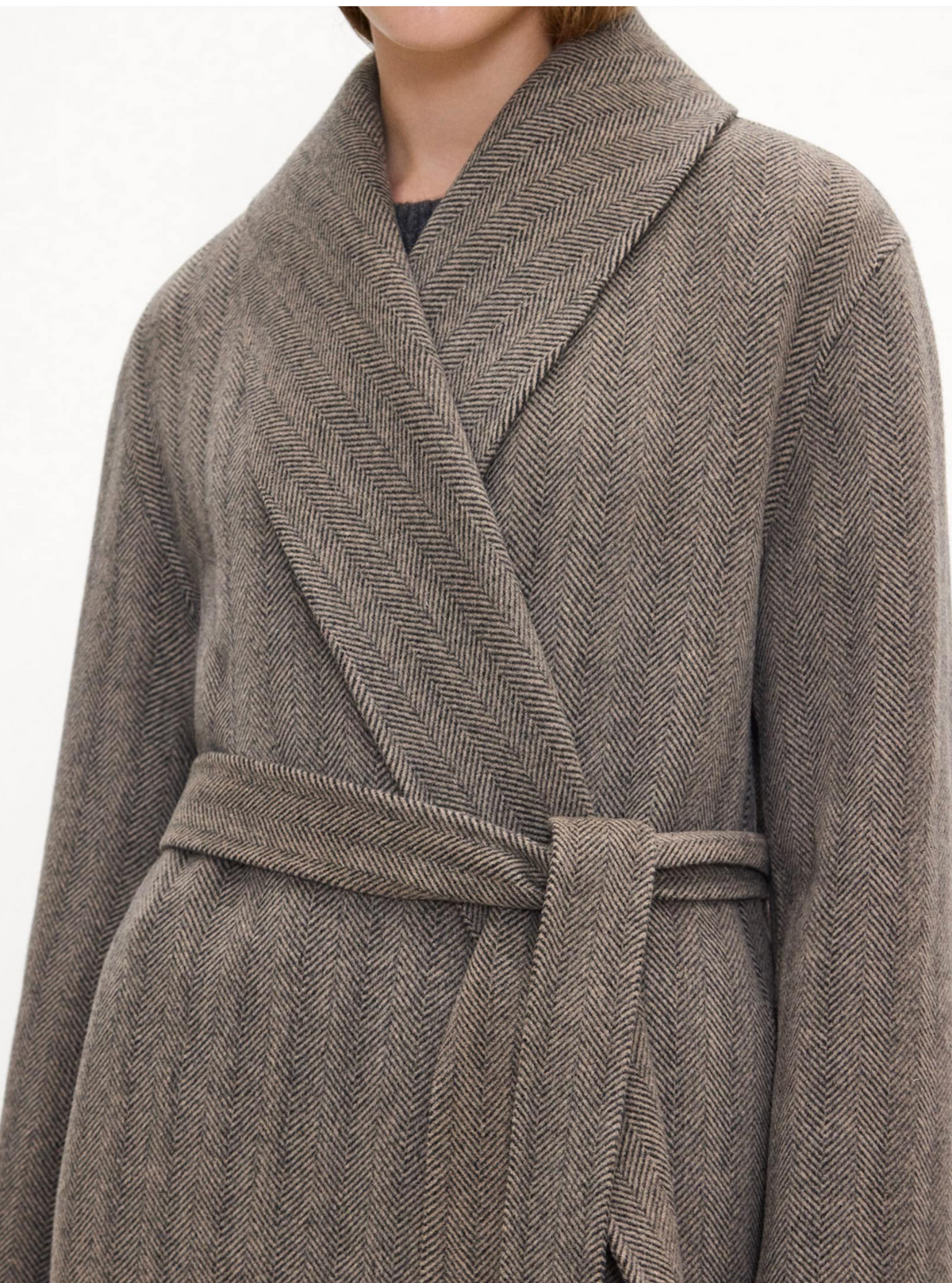 A close-up of a person wearing a taupe-brown herringbone patterned wrap coat with a matching tie belt, shown against a plain light background.