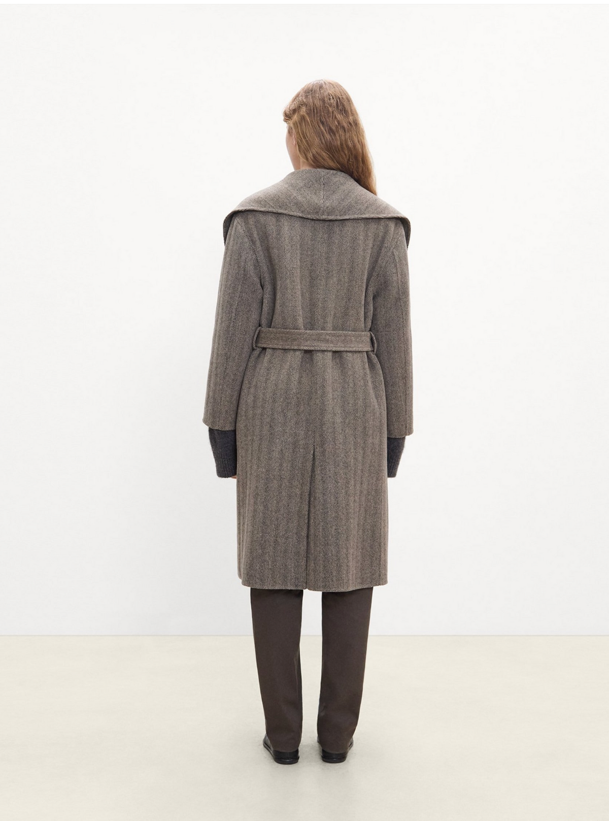 A model viewed from behind wearing a long, wide-collar coat in a textured taupe-brown and gray herringbone pattern, paired with dark brown trousers and black shoes, set against a plain white studio background.