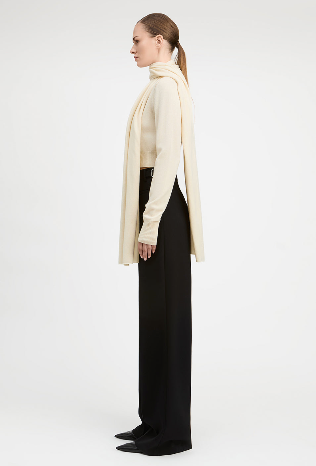 A model is shown in profile wearing a cream-colored knit turtleneck sweater with a long draped scarf, paired with black wide-leg trousers and black pointed-toe flats, against a plain white studio background.