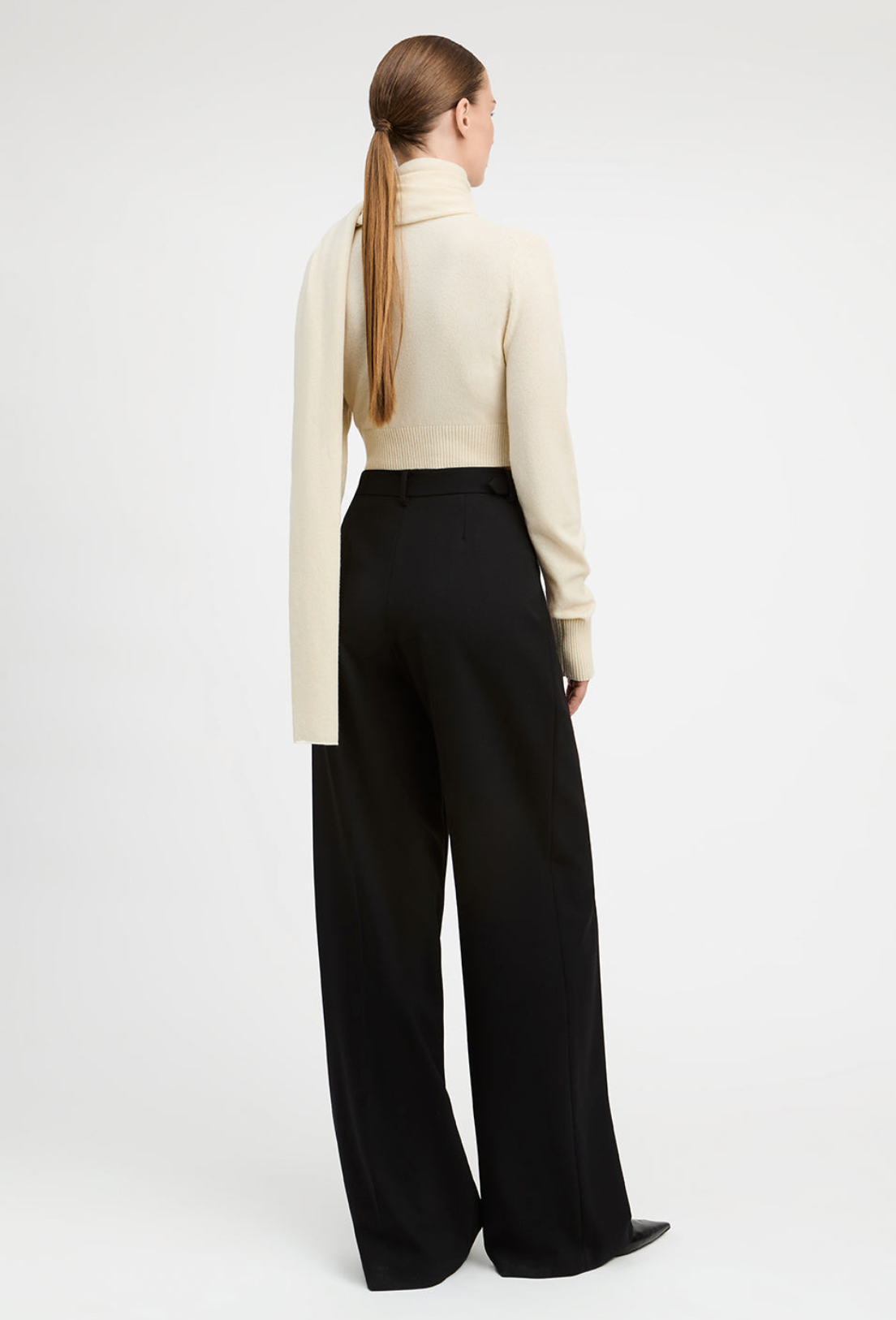 A woman shown from the back wearing a cream turtleneck sweater and black wide-leg trousers, photographed against a clean white studio background.