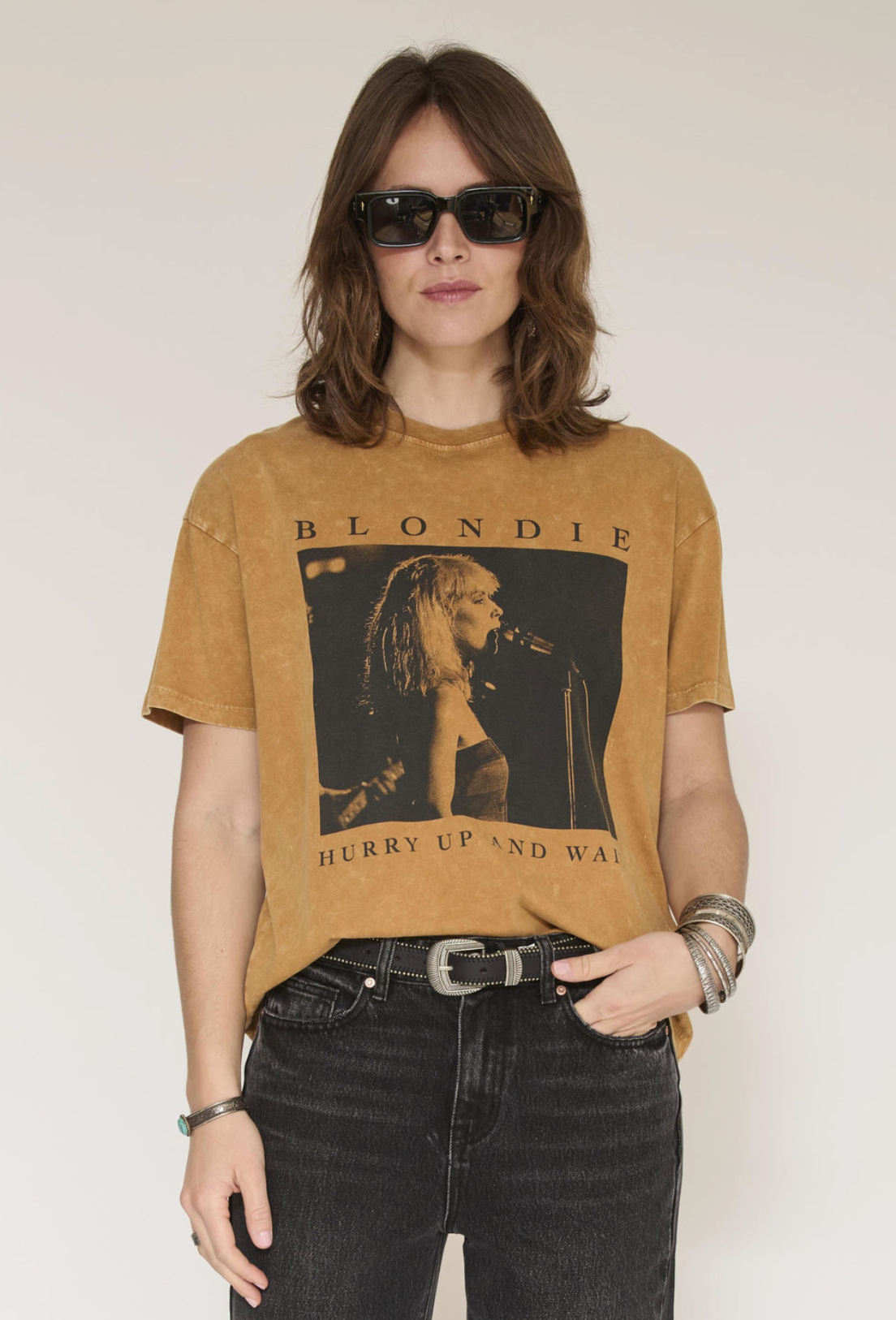 A woman modeling a cropped, faded tan t-shirt with a black graphic of Blondie performing and the text 'BLONDIE' and 'HURRY UP AND WALK' printed on the front. She is wearing black jeans, a silver belt, and sunglasses against a plain off-white background.