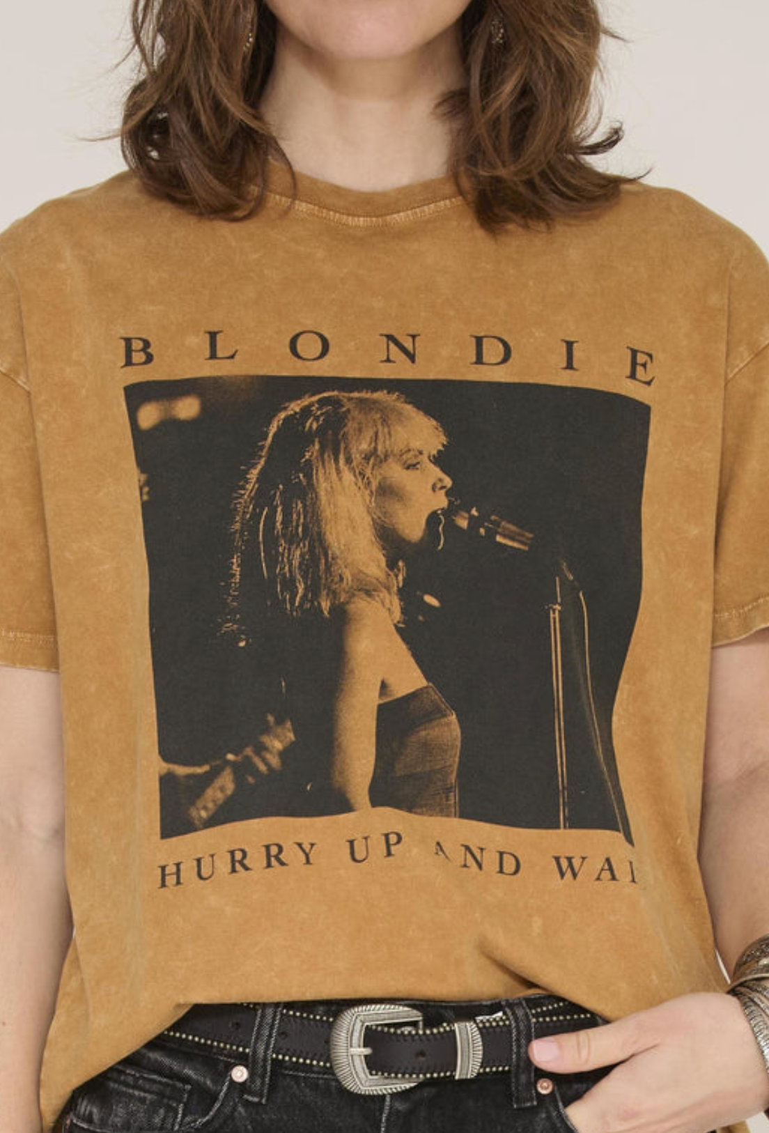 A person wearing a cropped, faded tan Blondie graphic T-shirt with a large black-and-tan band photo print on the front, paired with dark jeans and a silver belt.