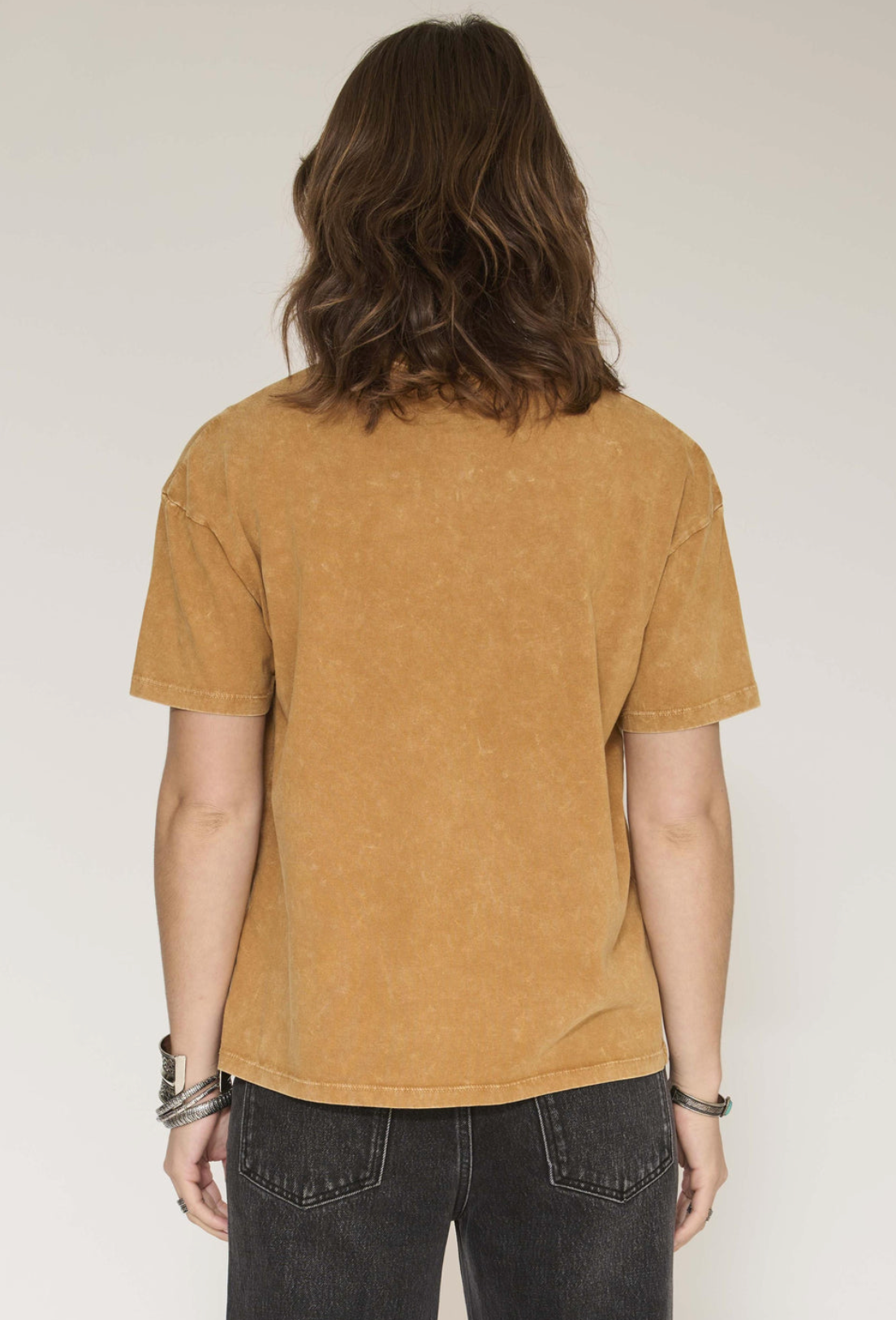 A person viewed from the back wearing a short-sleeve cropped t-shirt in a washed mustard yellow color, paired with dark gray jeans, against a plain light gray studio background.