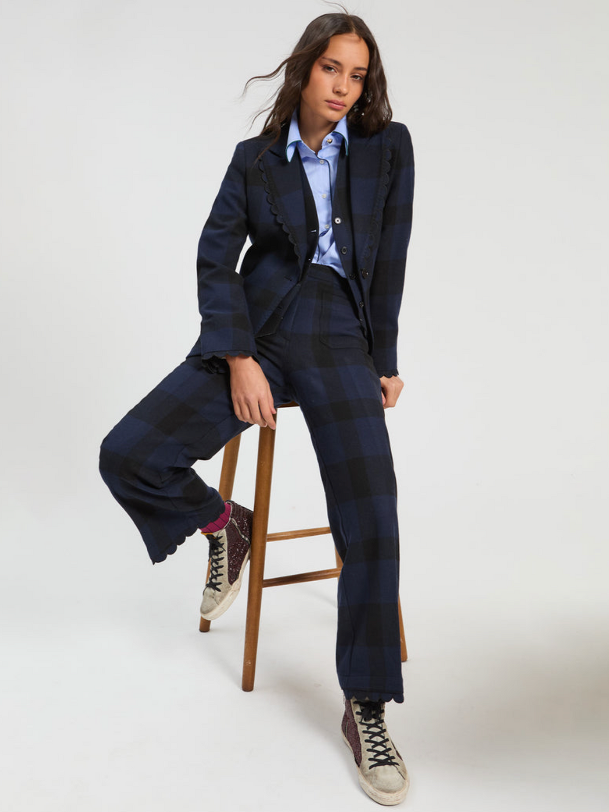 A model wearing a dark navy blue plaid blazer and matching wide-leg trousers, styled with a light blue button-down shirt and dark sneakers, sitting on a wooden stool against a light gray studio background.
