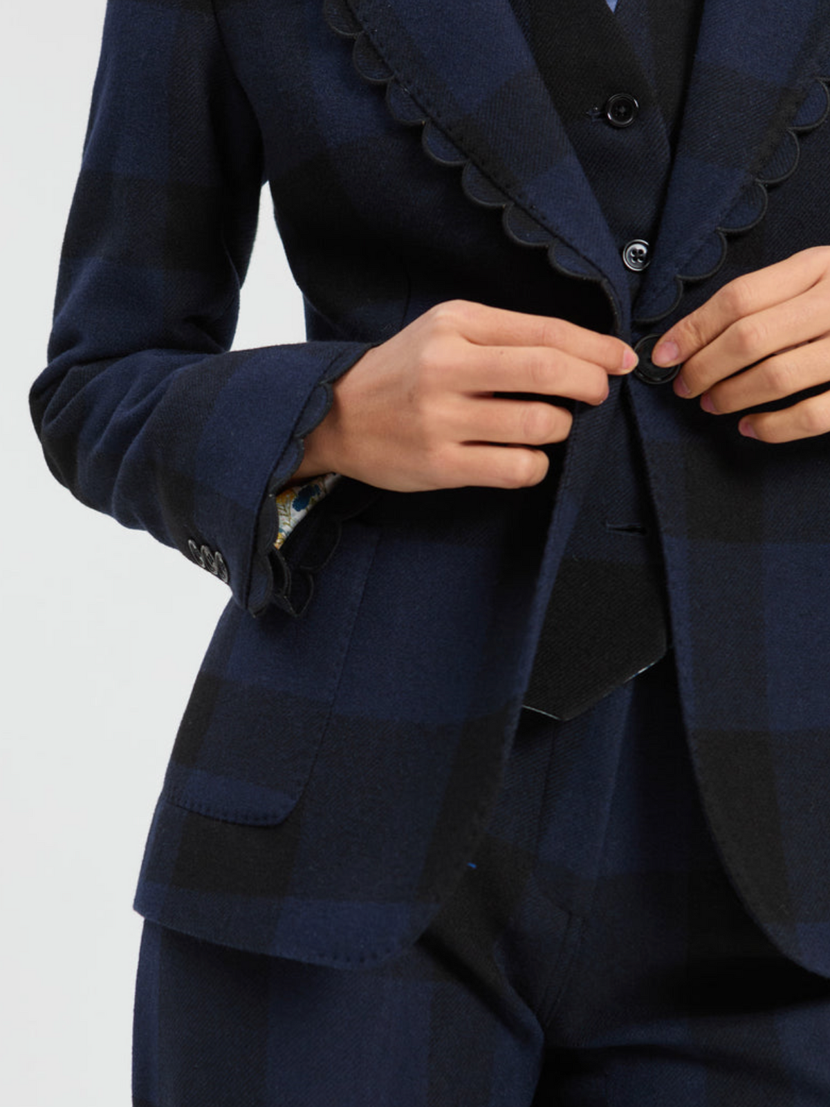 Close-up of a person wearing a navy and black plaid blazer with scalloped lapels, buttoning the front. The matching trousers are also visible against a plain light gray background.