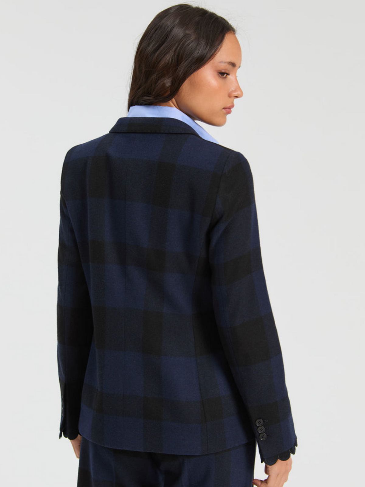 A woman shown from the back wearing a navy and black plaid blazer over a light blue collared shirt, against a plain light gray studio background.