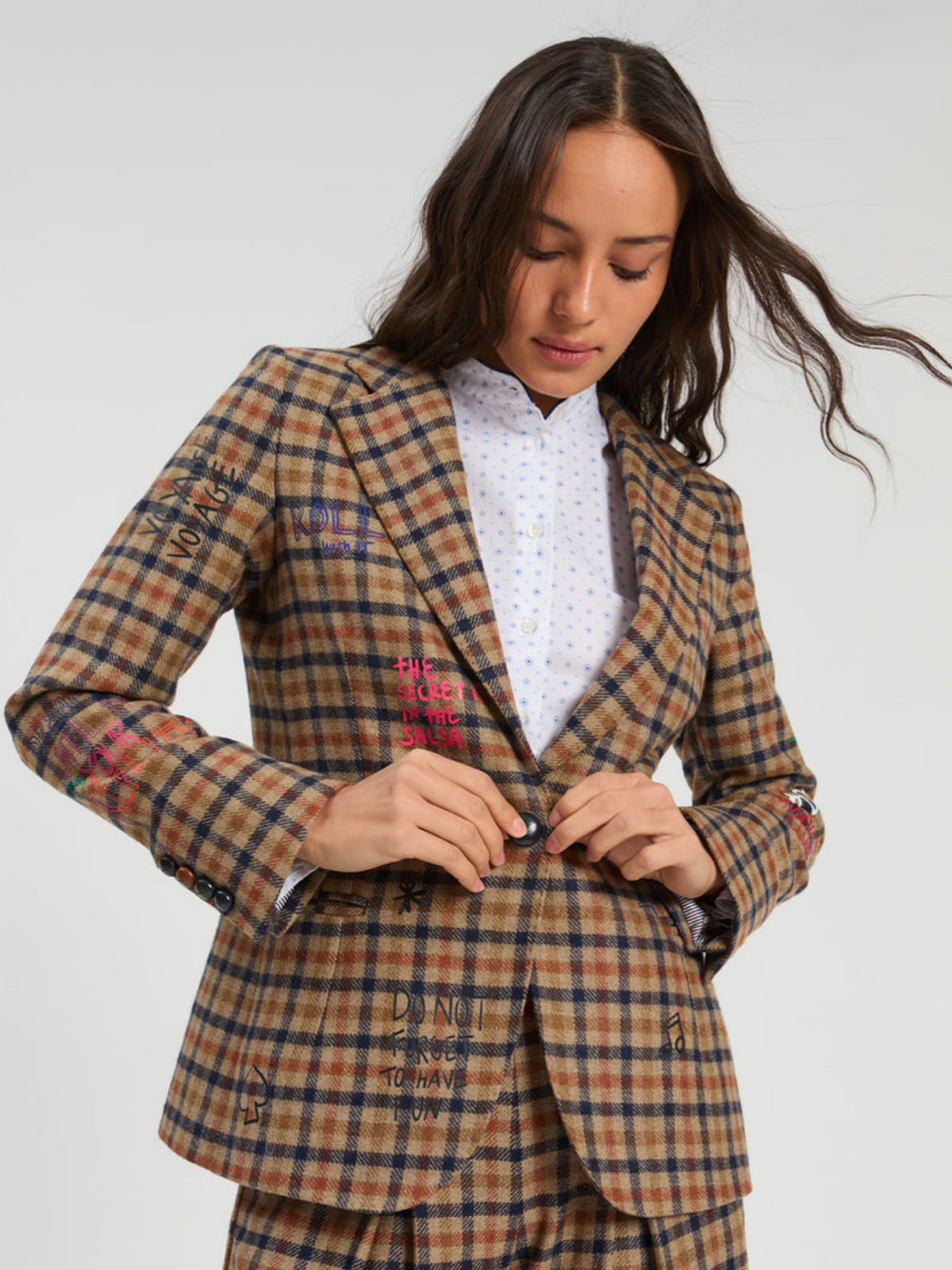 A woman wearing a brown and beige plaid blazer with colorful embroidered text details, styled over a white patterned shirt, against a light gray studio background.