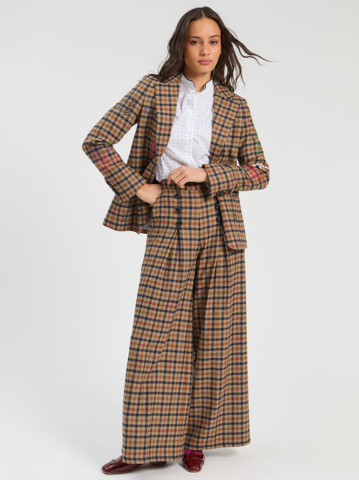 A model wearing a matching brown and beige plaid suit set, featuring a cropped blazer with red plaid details on the sleeves, wide-leg trousers, a white patterned shirt, and burgundy shoes, against a plain light gray background.