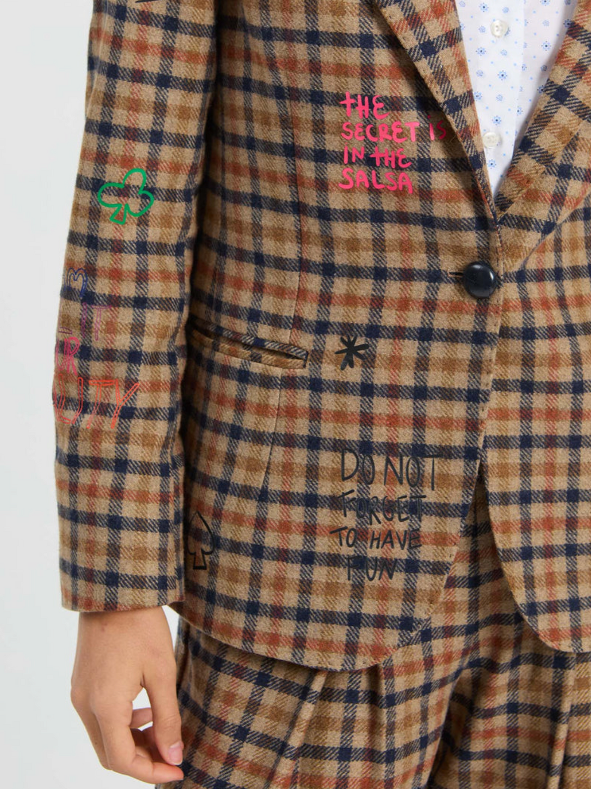 A close-up of a person wearing a brown, beige, and navy plaid blazer with colorful graffiti-style embroidered text and symbols, paired with matching trousers.