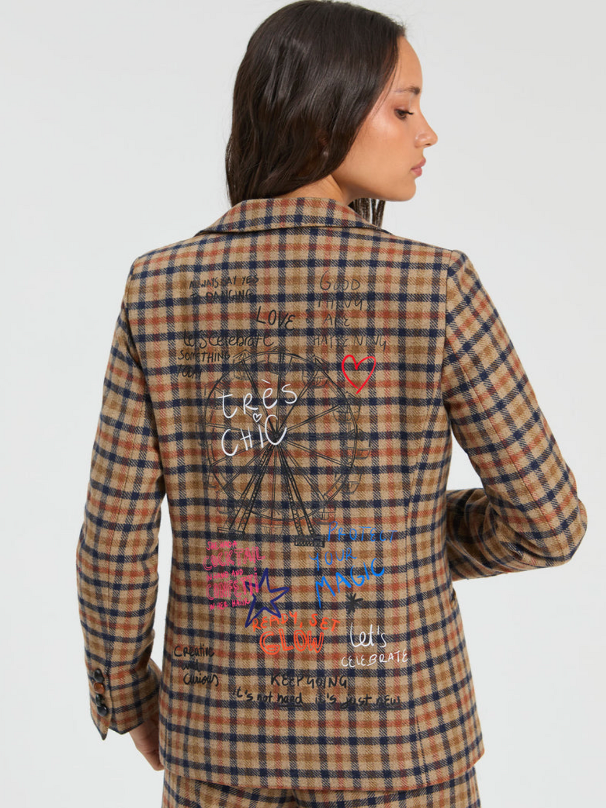 A woman shown from the back wearing a brown and navy plaid blazer with a large graphic print on the back. The print features a compass design with the words 'trees or CHIC' in white script, surrounded by various colorful text elements in red, pink, and blue.
