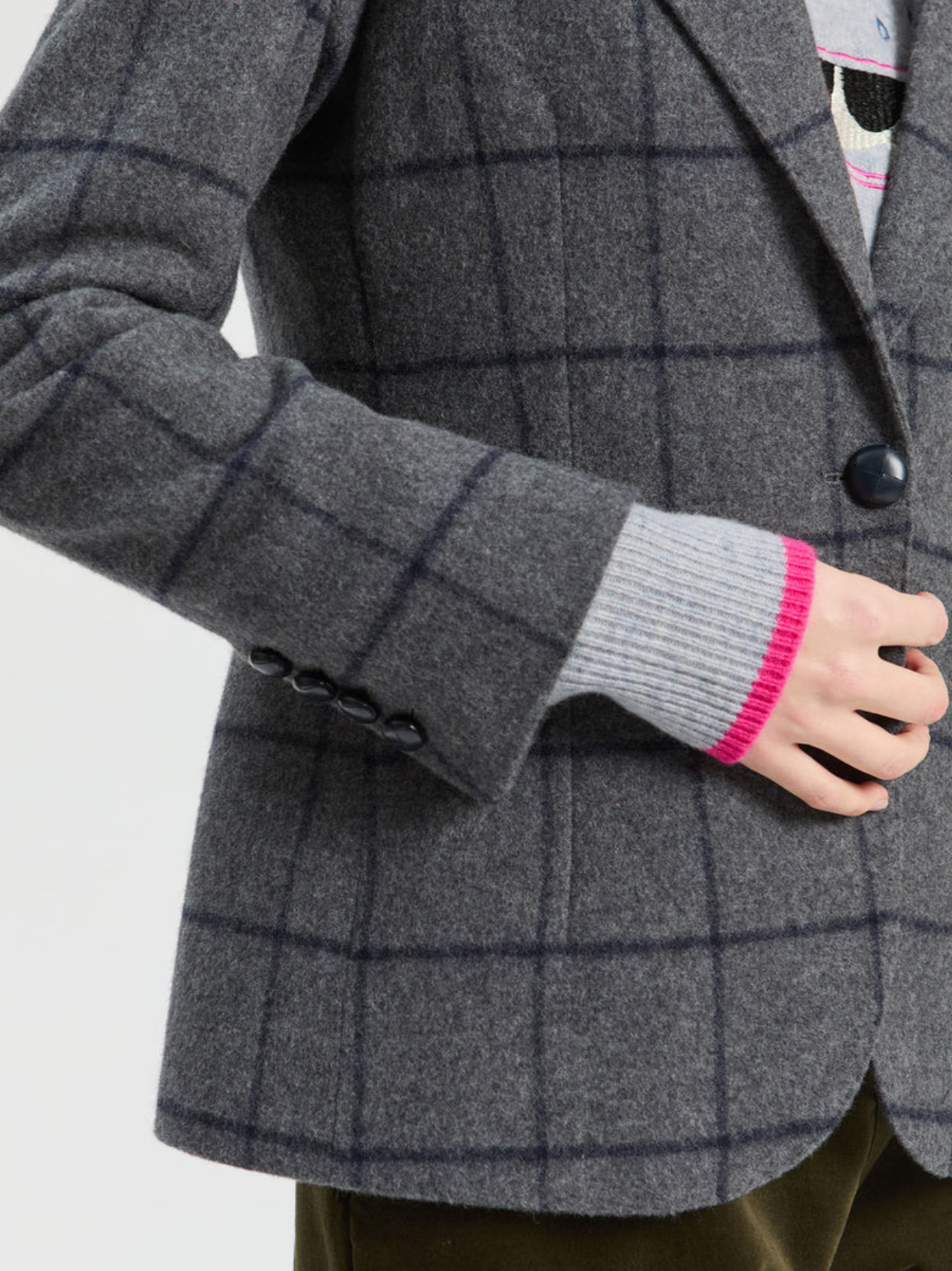 Close-up of a person wearing a gray wool-blend blazer with a dark windowpane plaid pattern, layered over a gray knit sweater with a bright pink cuff, and olive green trousers.