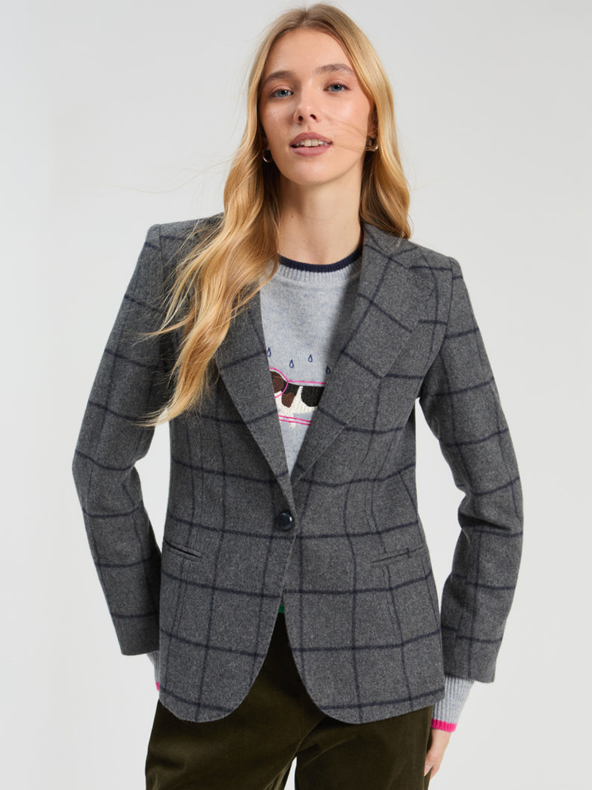 A woman modeling a gray windowpane plaid blazer over a gray graphic crewneck sweater and dark olive pants, photographed against a light gray studio background.