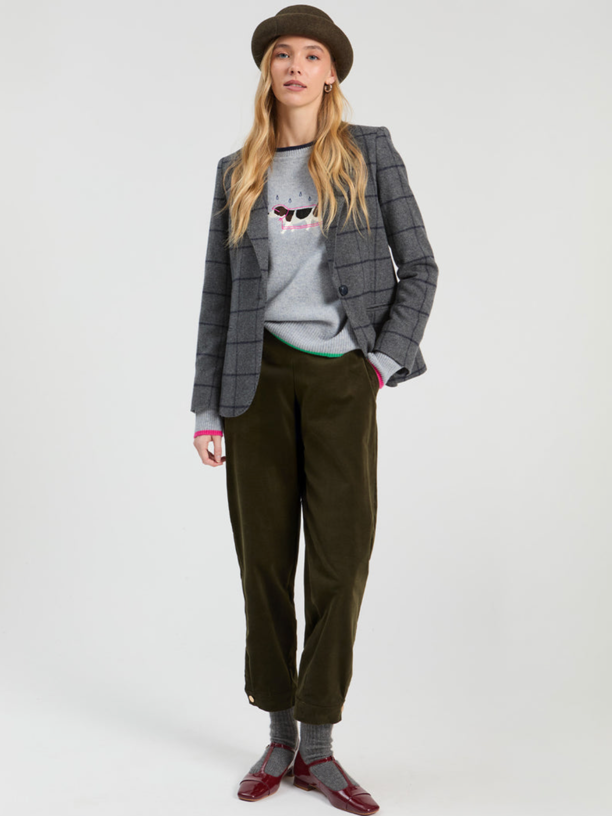 A model stands against a light gray studio background wearing a dark brown flat hat, a gray plaid blazer over a gray graphic sweatshirt, olive green corduroy trousers, gray socks, and burgundy loafers.