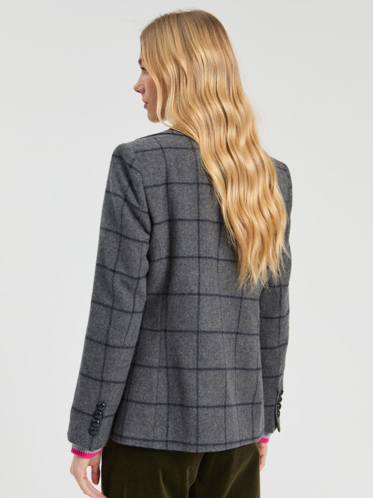 A woman with long wavy blonde hair is shown from the back wearing a gray windowpane plaid blazer and dark olive green trousers, photographed against a light gray studio background.