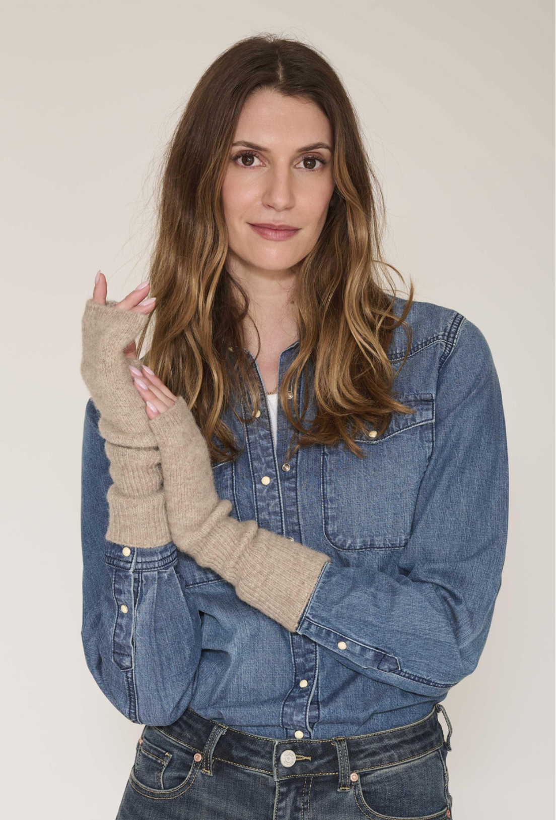 A woman wearing a pair of beige ribbed knit fingerless gloves with built-in wrist cuffs, styled over a blue denim shirt and jeans against a plain light background.