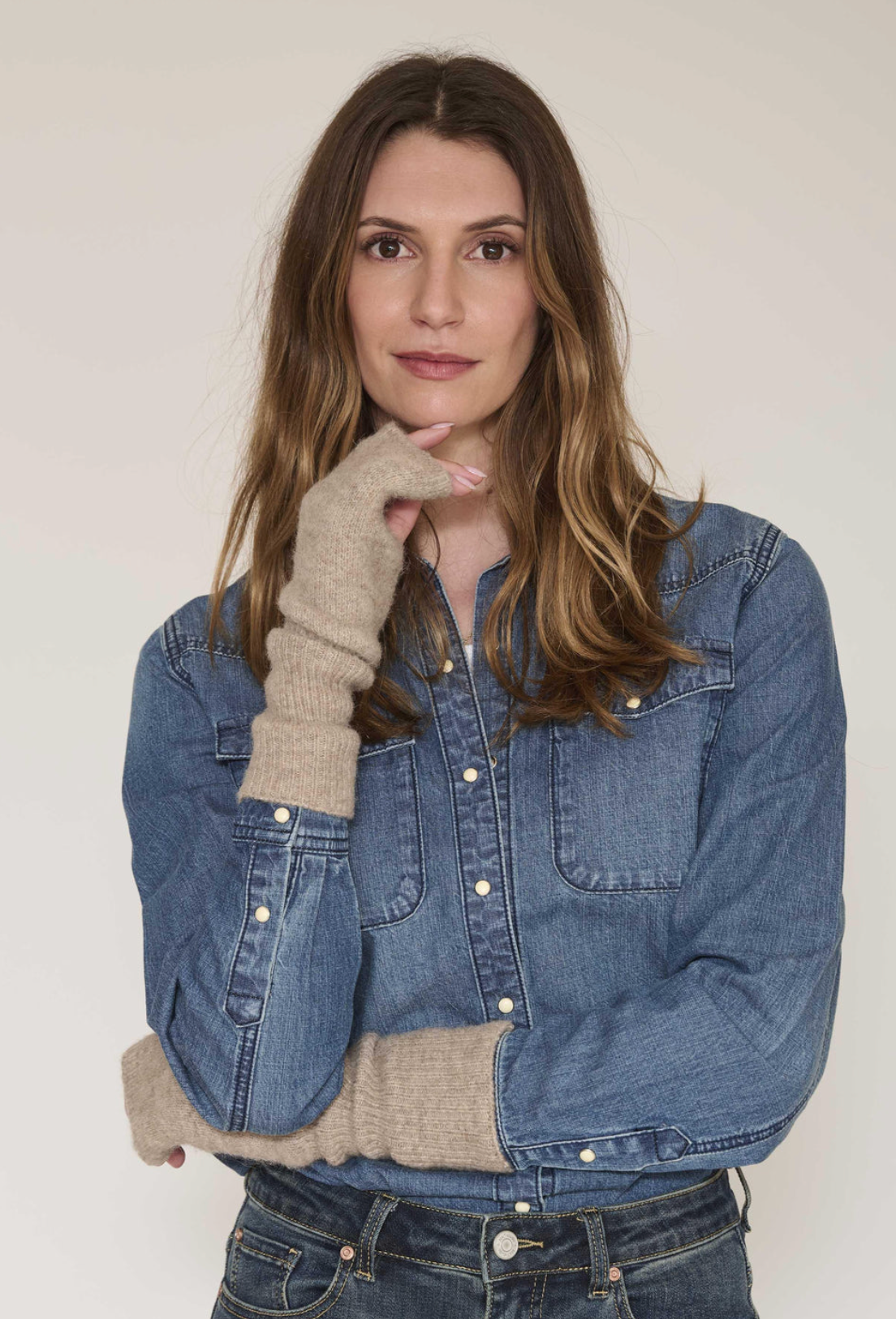 A woman wearing a blue denim jacket with beige knit thumbholes and a beige knit collar, paired with dark blue jeans, posing against a light off-white background.
