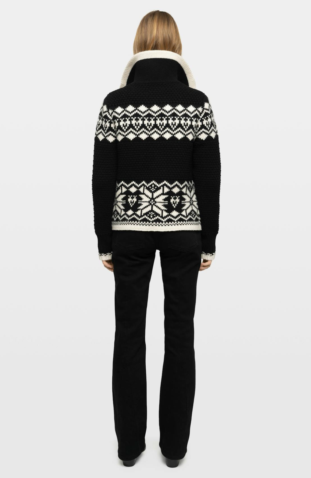 A model viewed from the back wearing a black knit sweater with a cream-colored geometric Fair Isle pattern across the shoulders and upper back, paired with black trousers, against a light gray studio background.