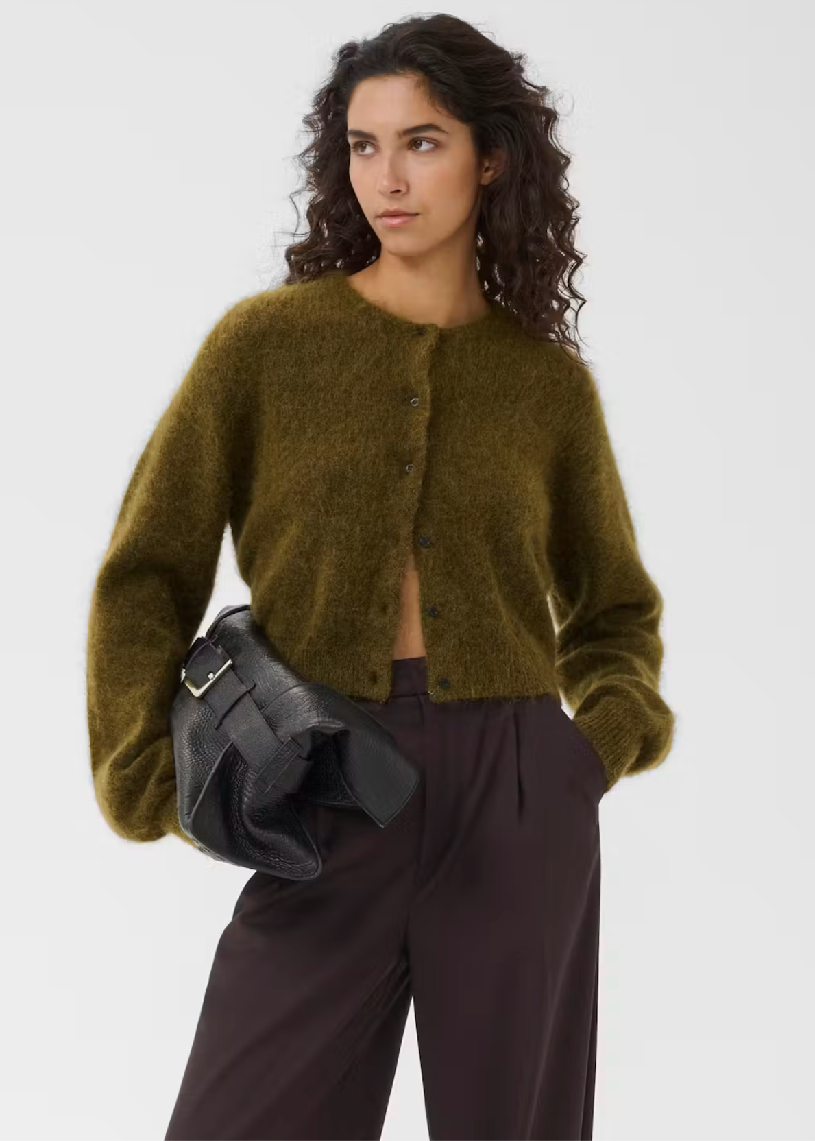 A woman models a fuzzy olive green cropped cardigan with front buttons, paired with dark brown trousers and a black leather clutch bag, against a plain light gray studio background.
